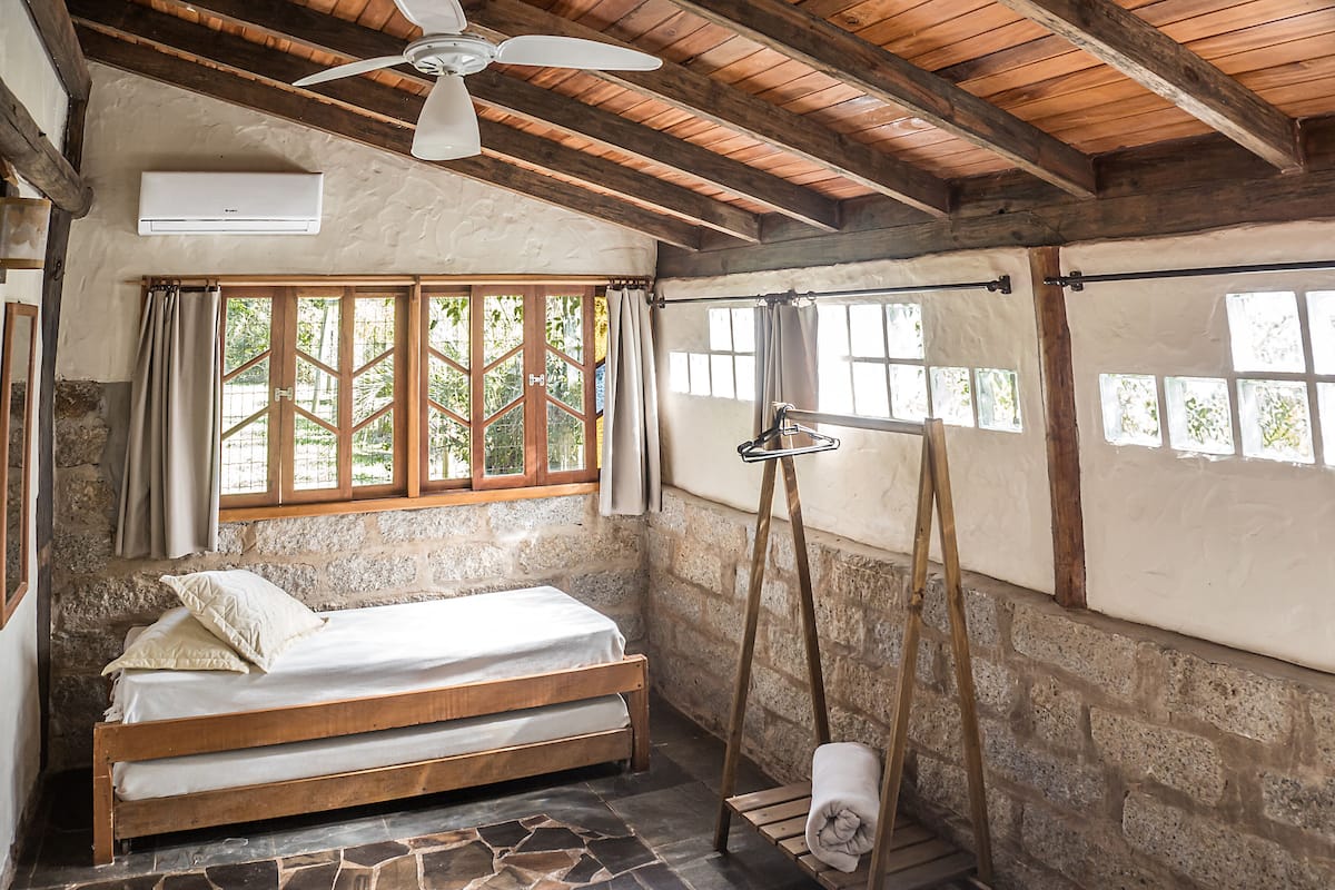 Ein rustikales Schlafzimmer mit einem hölzernen Bett, Steinwänden, einem Deckenventilator und großen Fenstern mit Vorhängen.