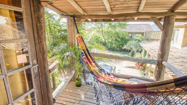 A hammock under a wooden structure with a view of a garden and a building in the background. - Pousada Morro das Palmeiras (Imbituba)