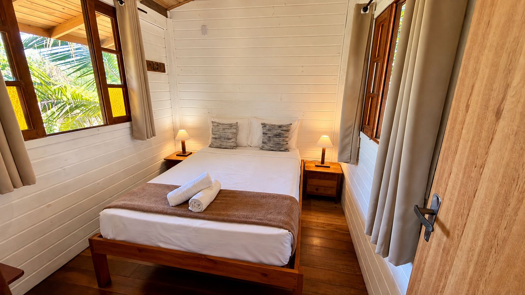 Una cama con estructura de madera, ropa de cama blanca y dos toallas enrolladas, con mesitas de noche con lámparas a cada lado, en una habitación con paredes de madera y una ventana con cortinas.