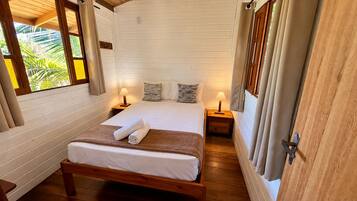 Una cama con estructura de madera, ropa de cama blanca y dos toallas enrolladas, con mesitas de noche con lámparas a cada lado, en una habitación con paredes de madera y una ventana con cortinas.