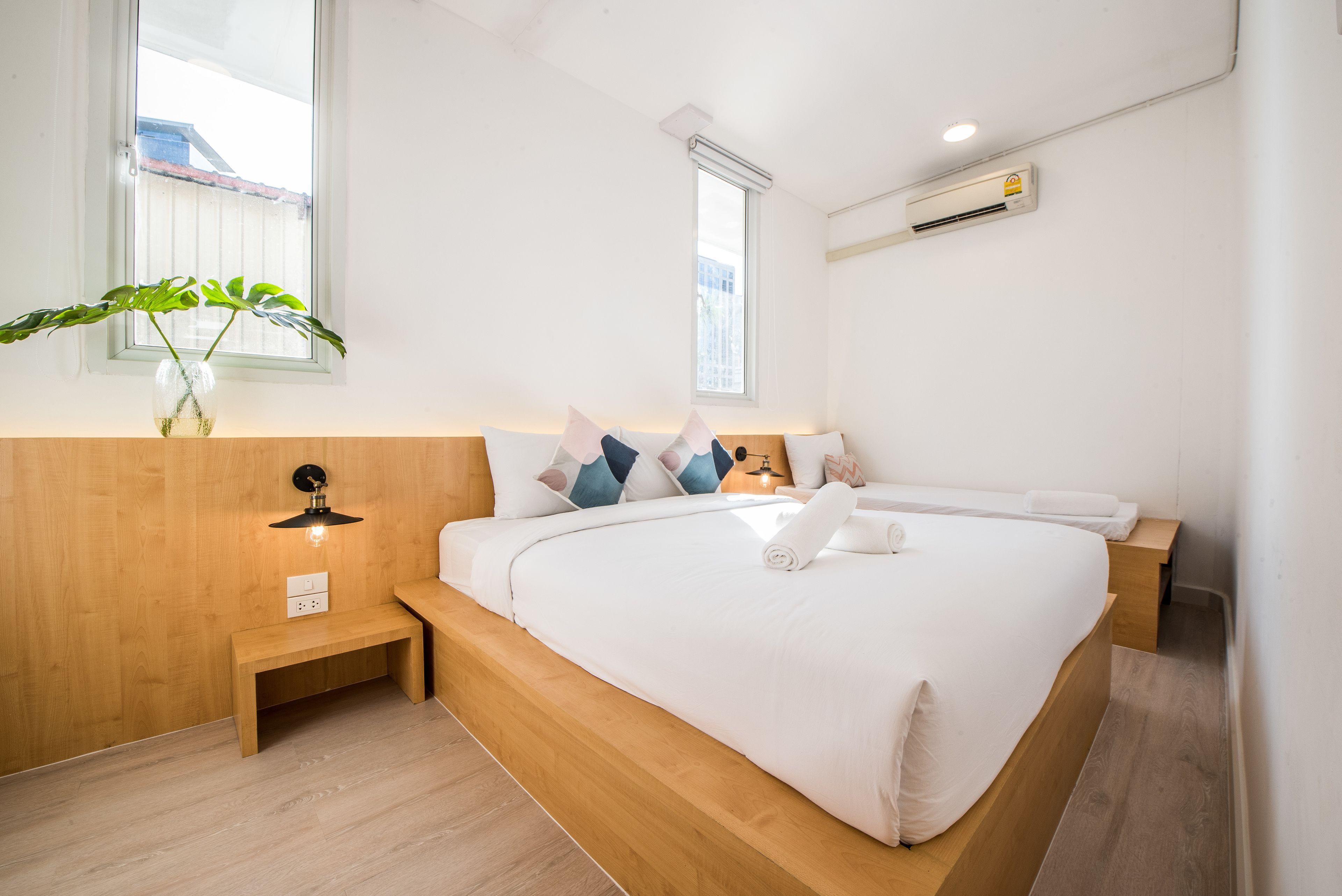 Una habitación de hotel moderna con cabecera de madera, una mesita de noche con lámpara, una cama grande con ropa blanca y una planta en maceta sobre el alféizar.
