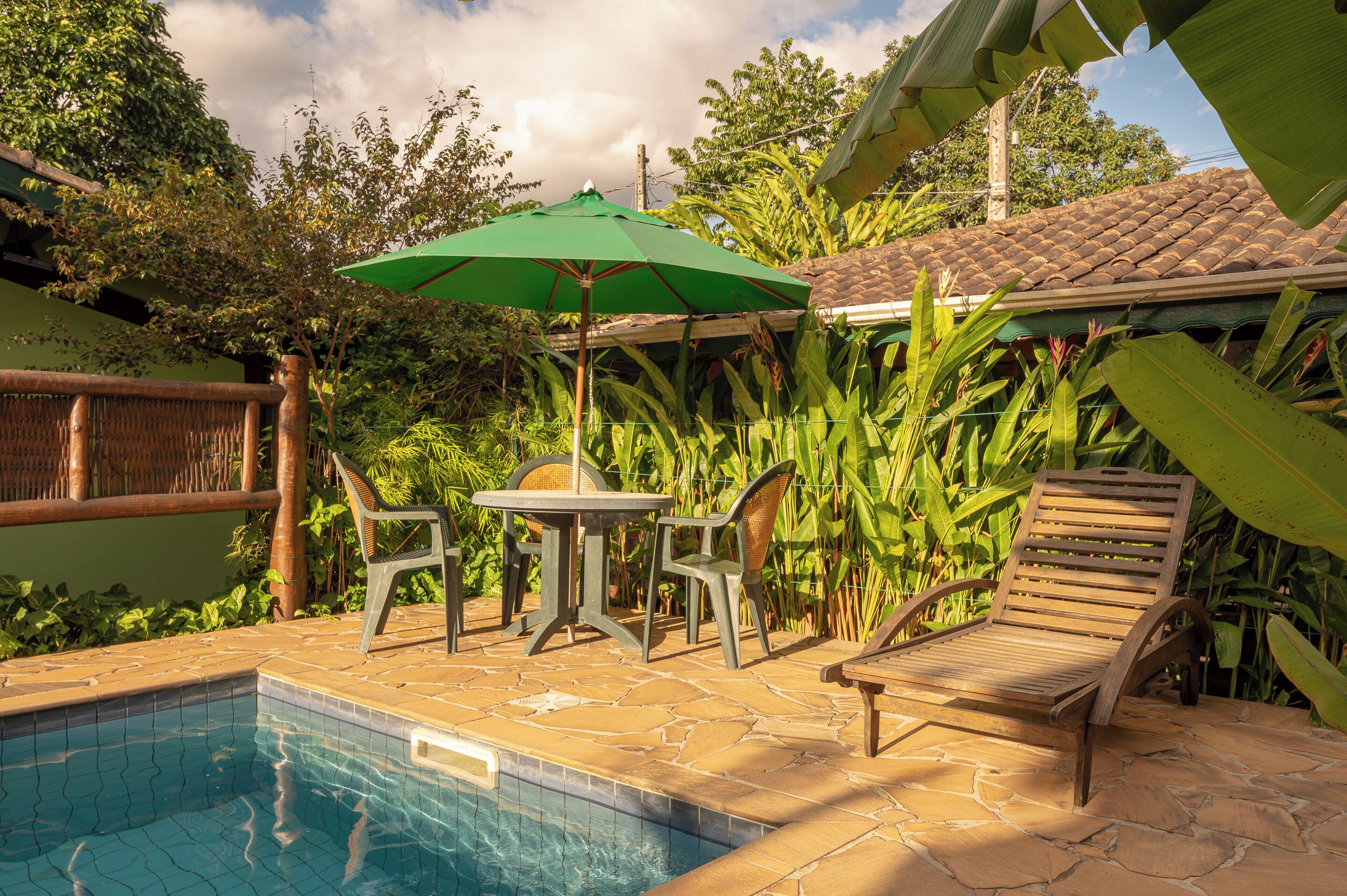 A pool area with a tiled floor, a table with chairs, a wooden lounge chair, and a green umbrella.