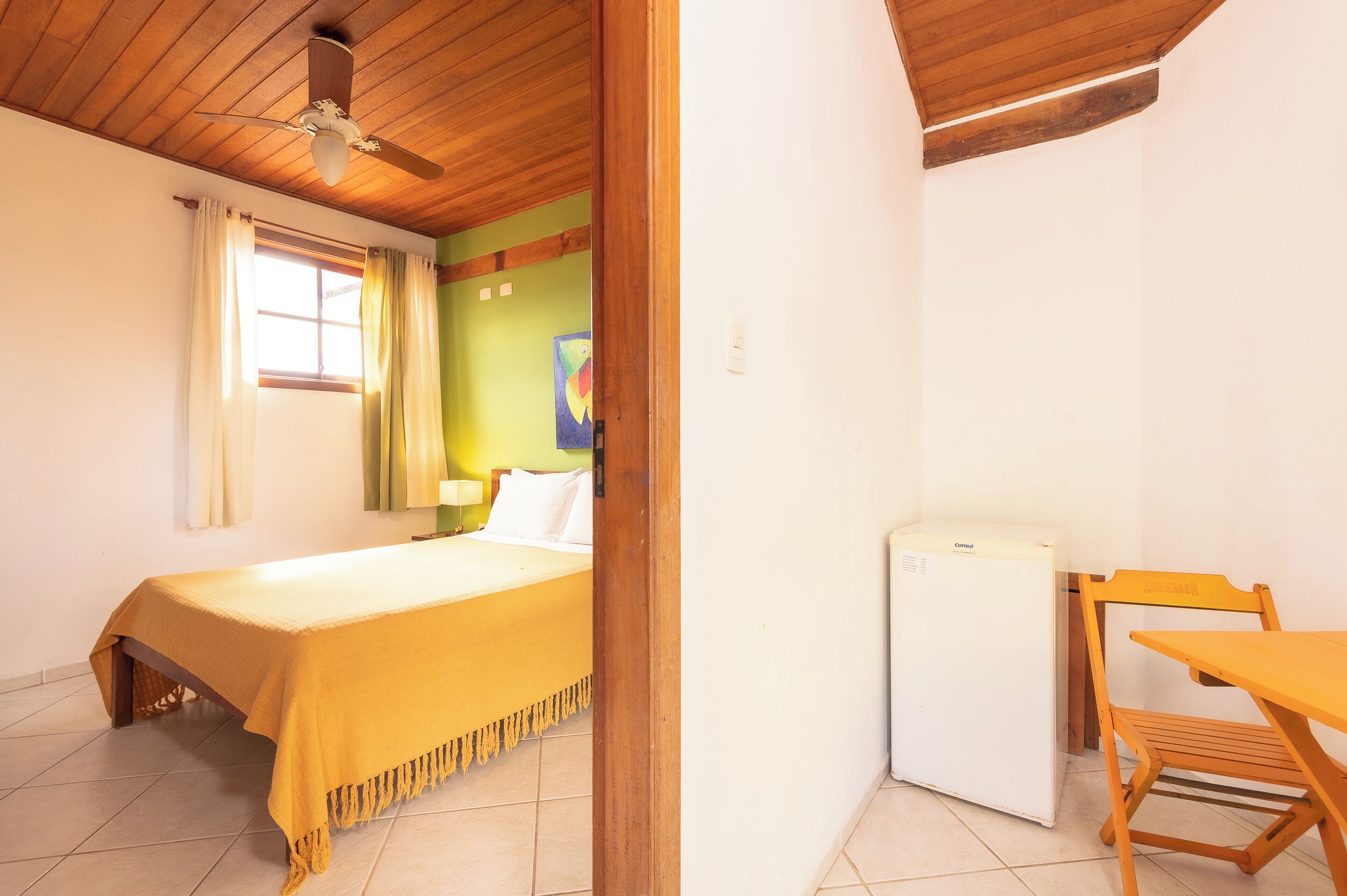 A room with a bed, a small table, a chair, a ceiling fan, and a refrigerator.