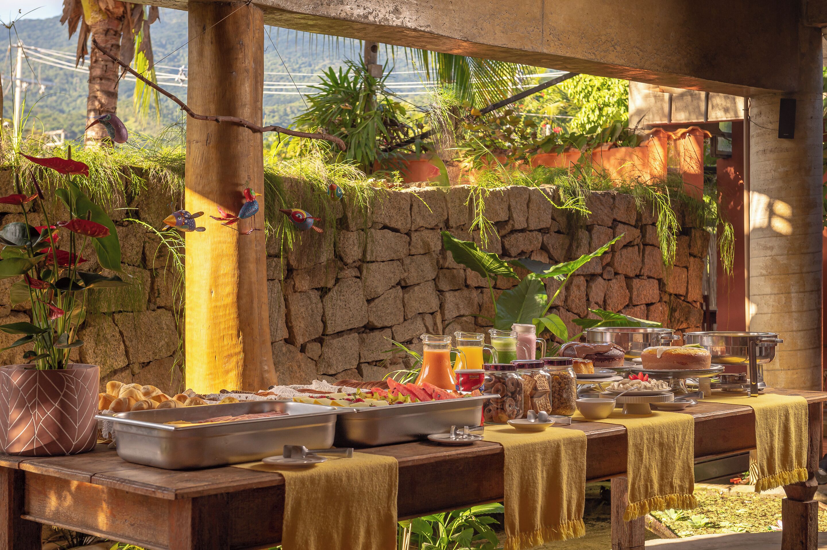 A buffet setup with various food items and drinks, set against a stone wall and surrounded by greenery.