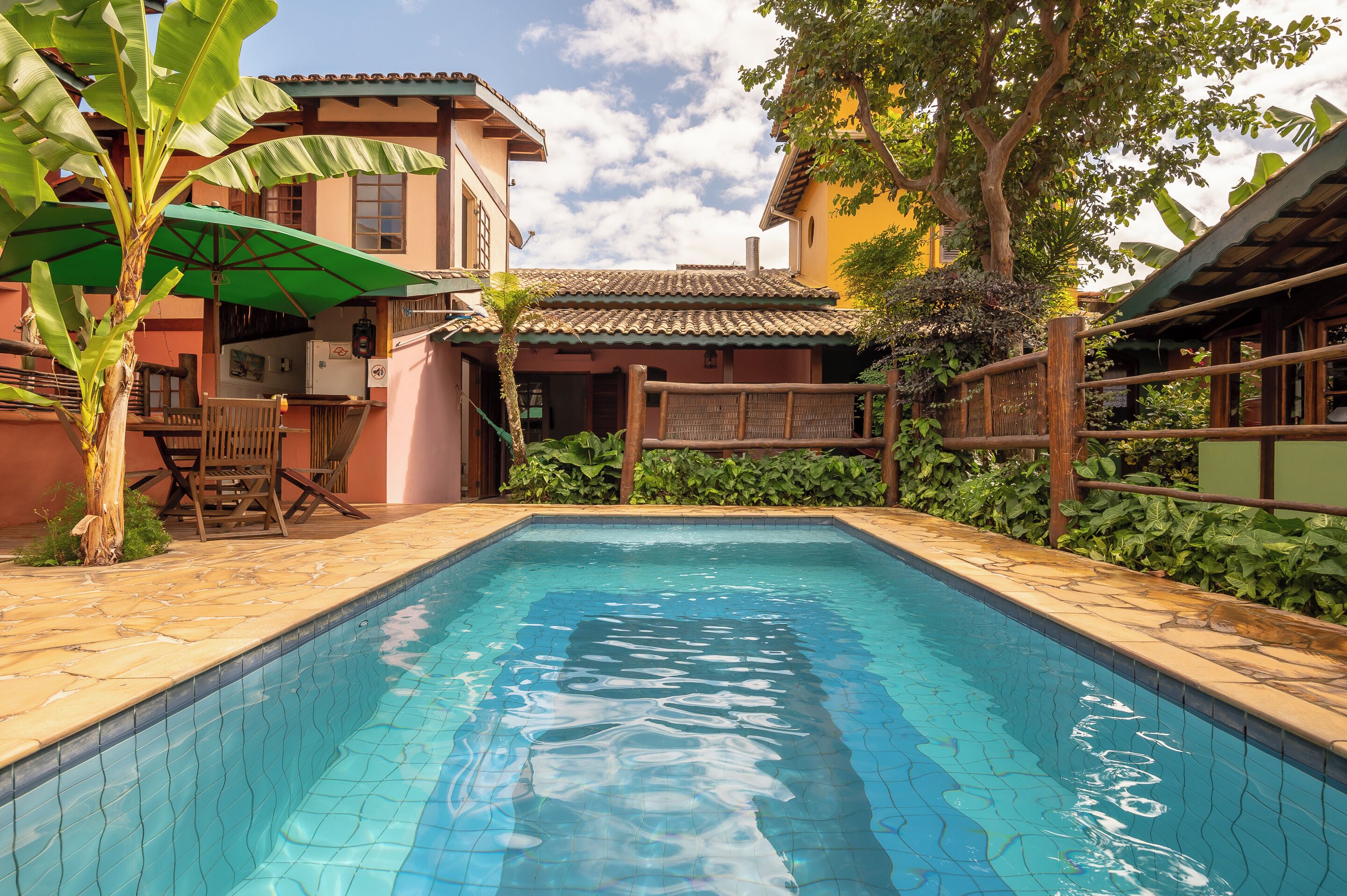 A swimming pool with tiled edges, surrounded by a patio area with a wooden fence, a wooden table, and chairs.