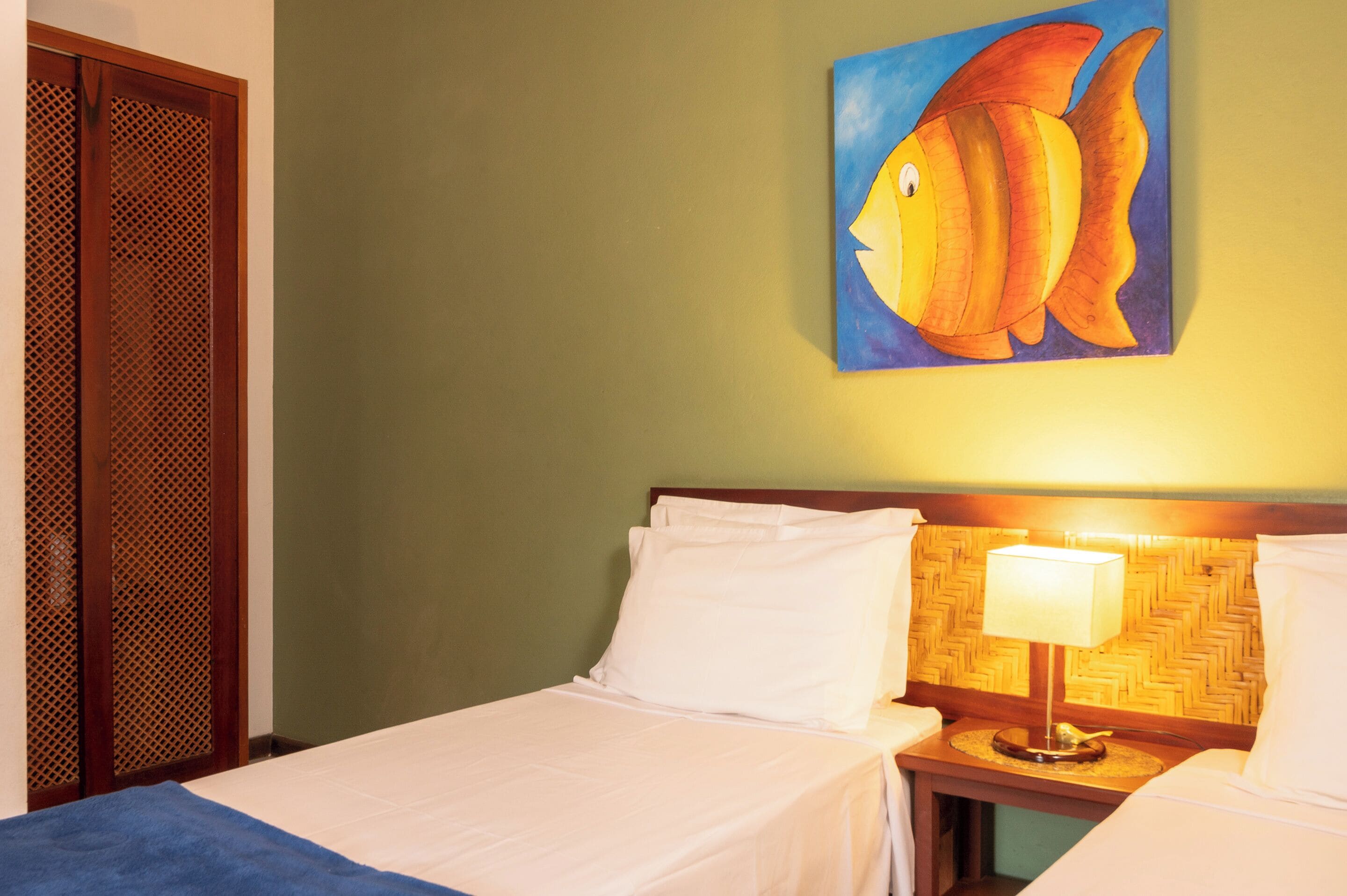 Ein Hotelzimmer mit einem Bett, einem Nachttisch mit Lampe, einem Wandbild von einem Fisch und einer Holztür mit Netzmuster.