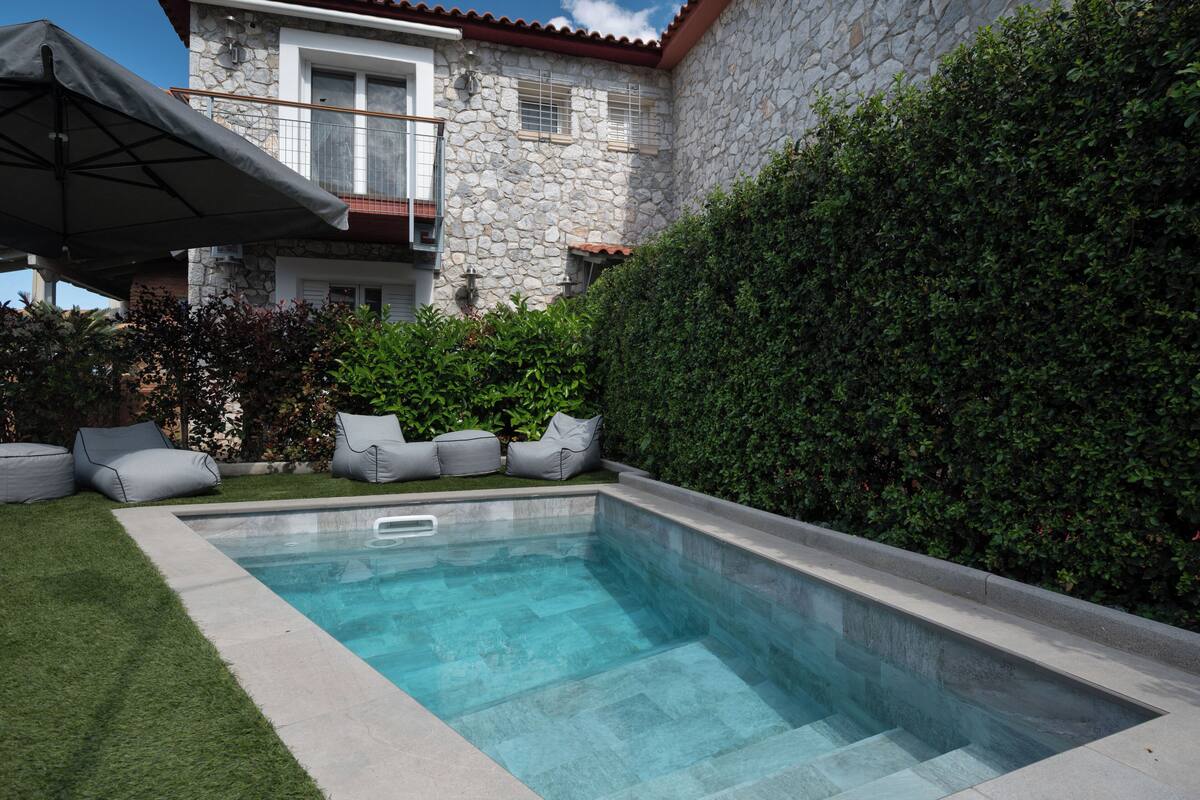 Una piscina con un muro de piedra y un seto, rodeada de césped y mobiliario de exterior.