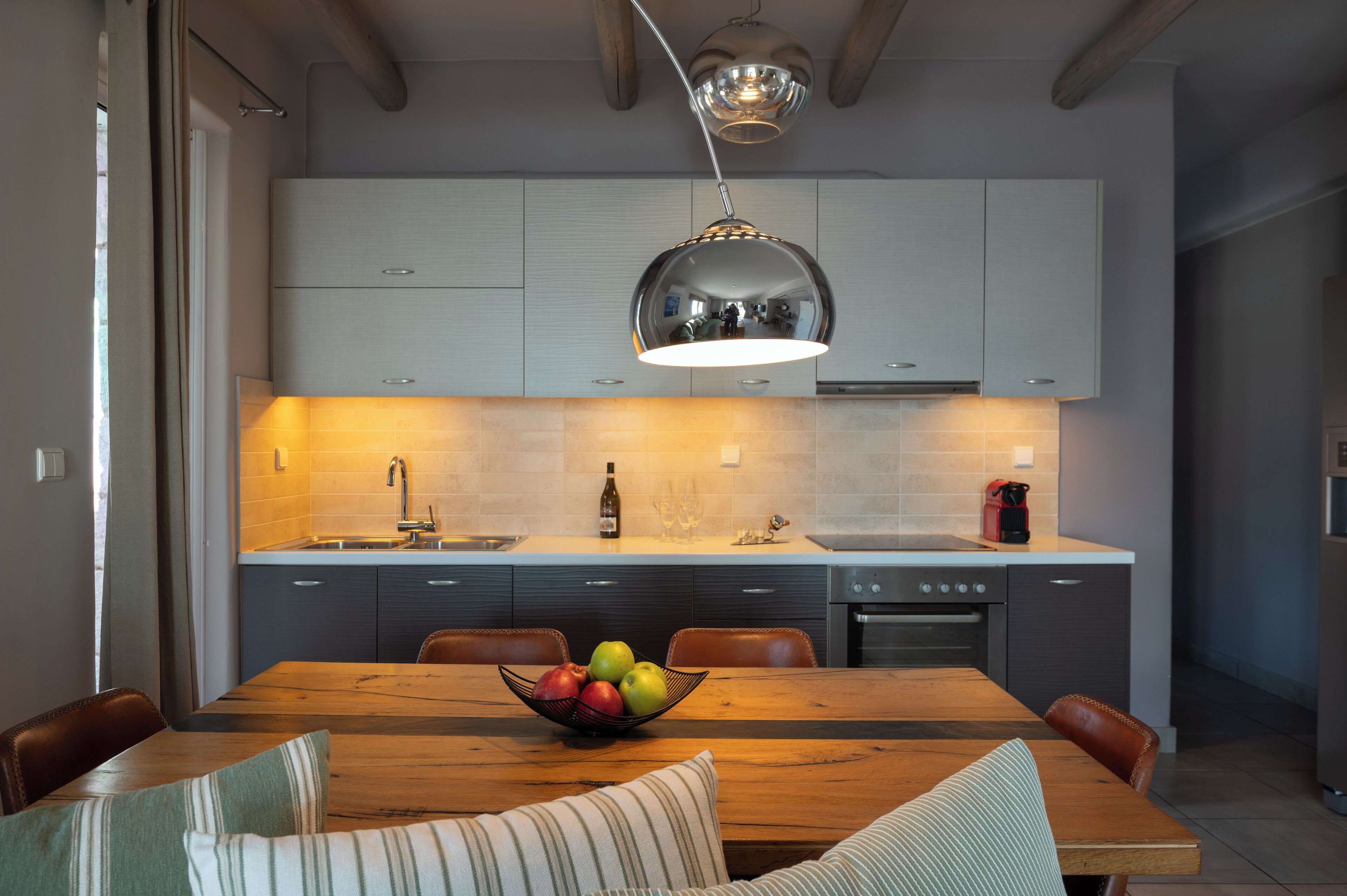 A modern kitchen with a wooden dining table, a bowl of fruit, and a pendant light.