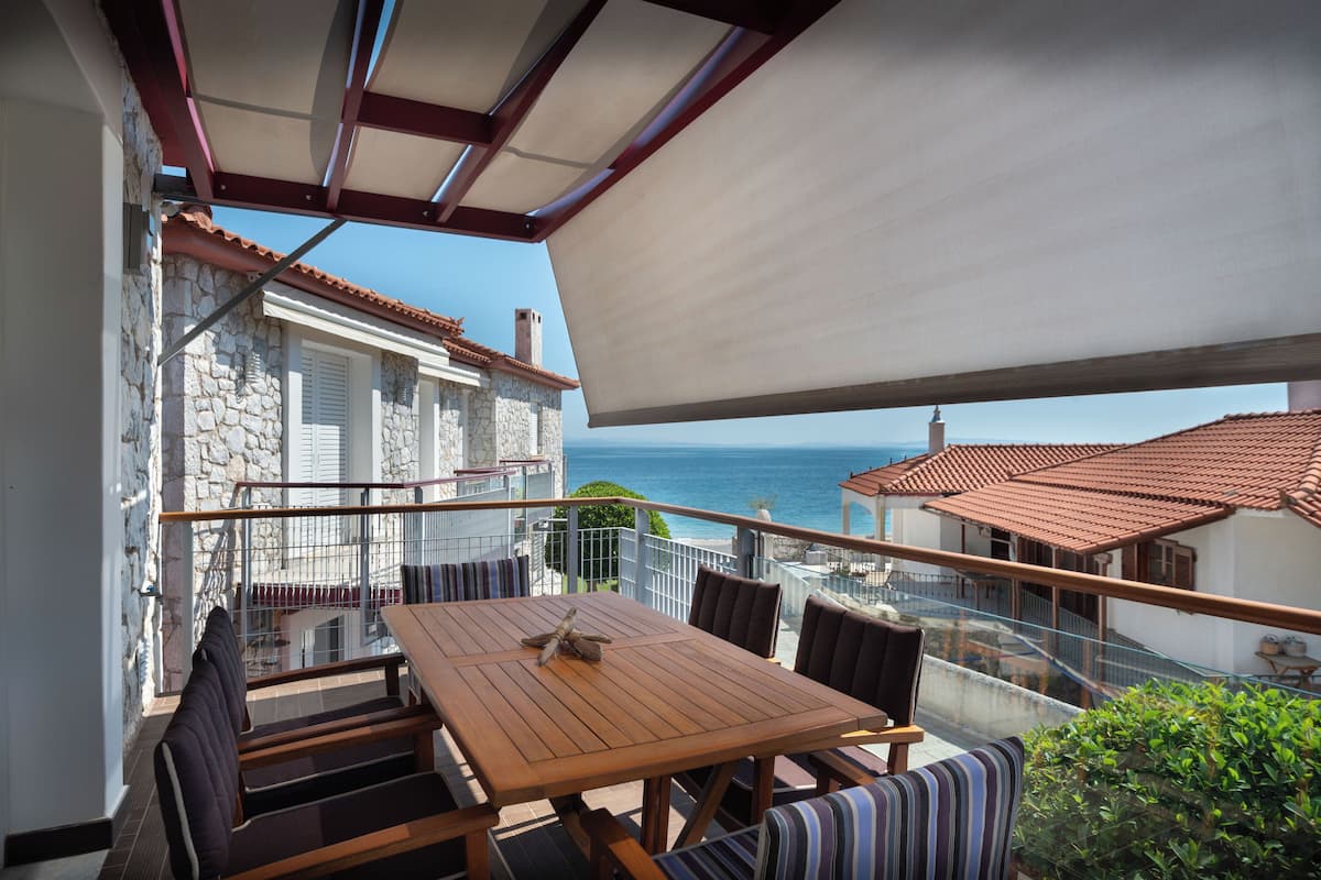 Un balcón con mesa y sillas de madera, un muro de piedra y vista al mar.