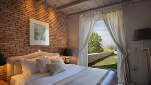 Un dormitorio con pared de ladrillo, cama con ropa de cama blanca, mesita de noche con lámpara y vista a un jardín por la ventana.