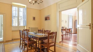Ein Esszimmer mit einem großen Holztisch, Stühlen und einer Kronleuchter. Es gibt Fenster mit Vorhängen, eine Heizung und eine Tür zu einem weiteren Raum.