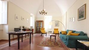 Ein großzügiger Wohnraum mit hoher Decke, einem Kronleuchter, einem grünen Sofa mit gelben Kissen, einem Holzschrank und einem gemusterten Teppich.