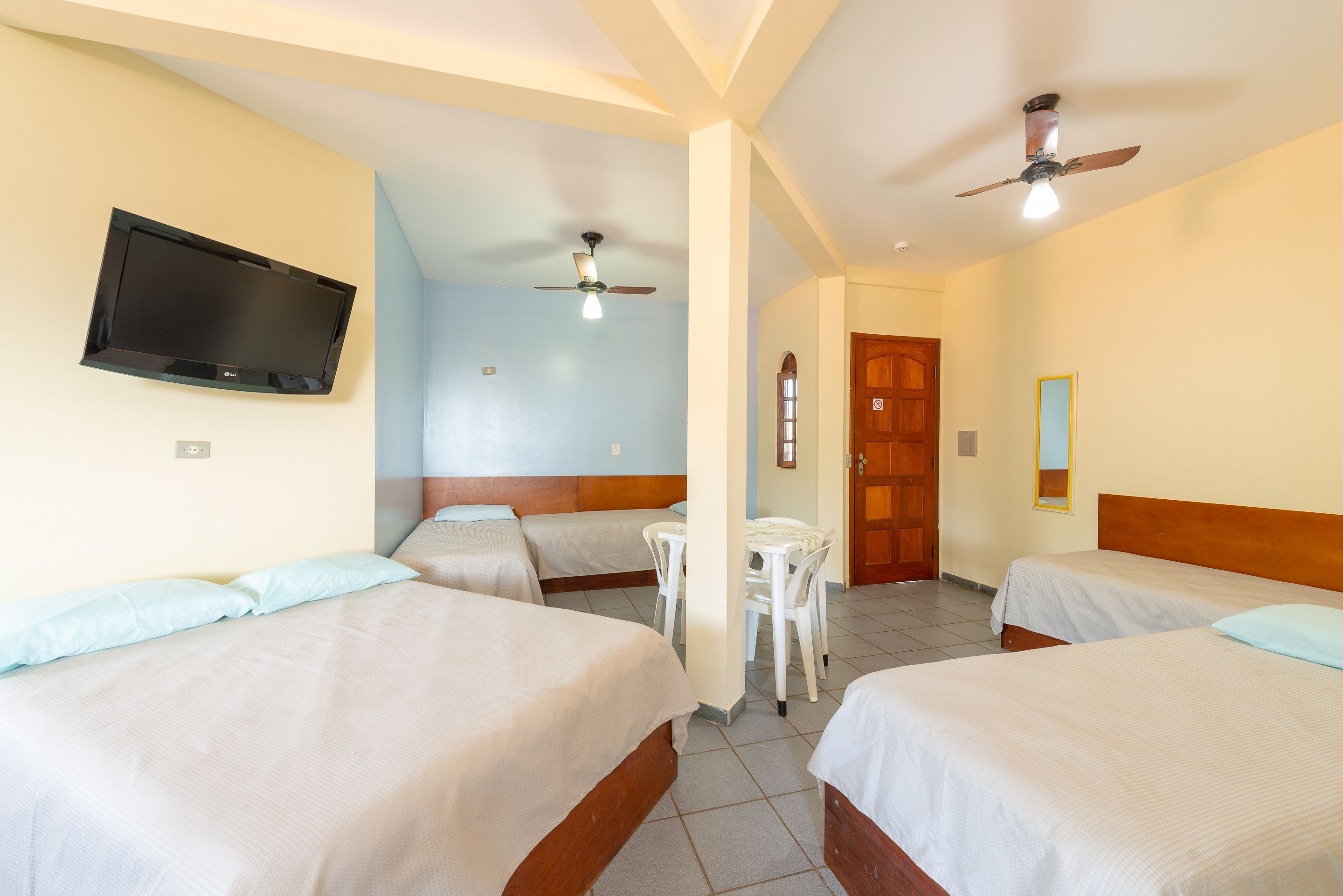 A hotel room with two beds, a TV mounted on the wall, a ceiling fan, and a small table with chairs.