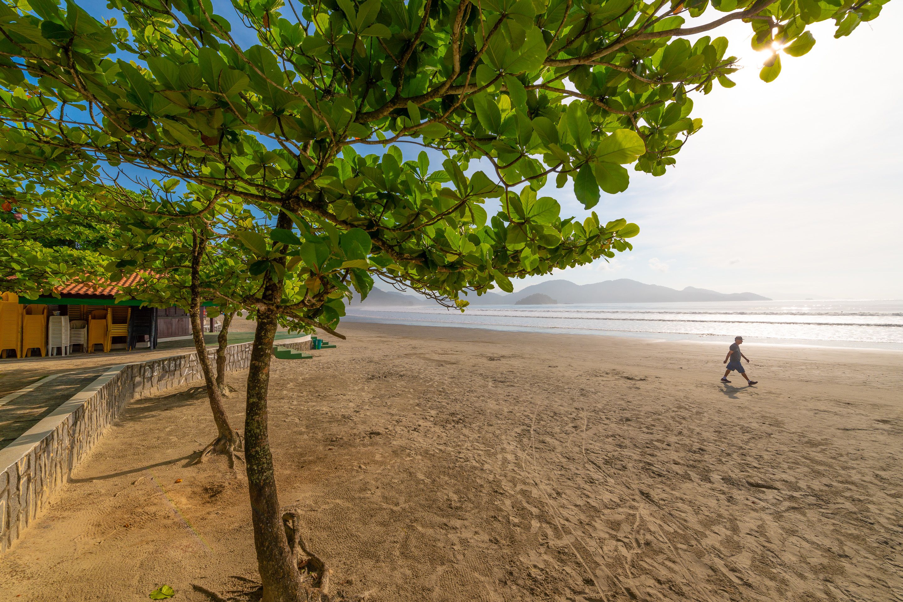 Une plage où l’on voit une personne qui se promène, des arbres et un bâtiment avec un toit en tuiles.