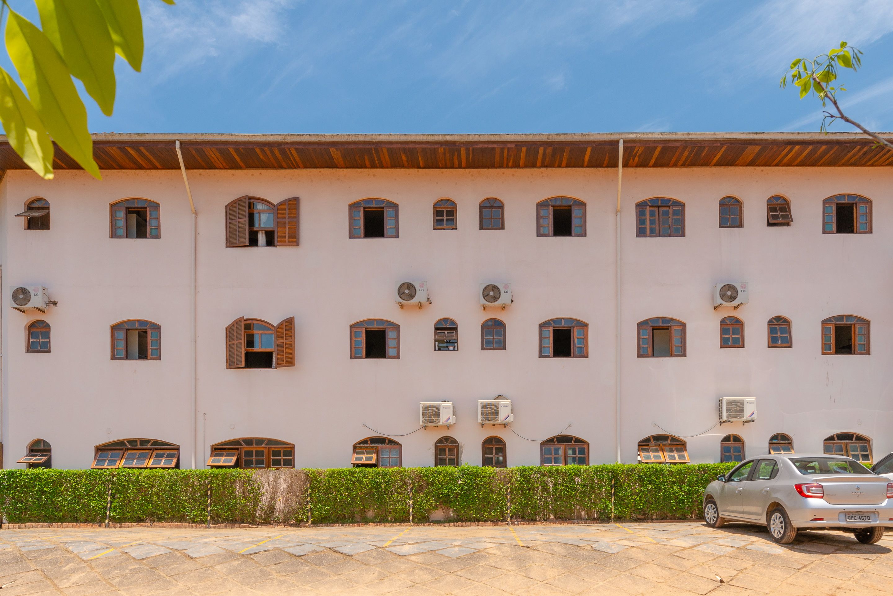 A building with multiple arched windows and air conditioning units, a paved parking area, and a hedge in the foreground.