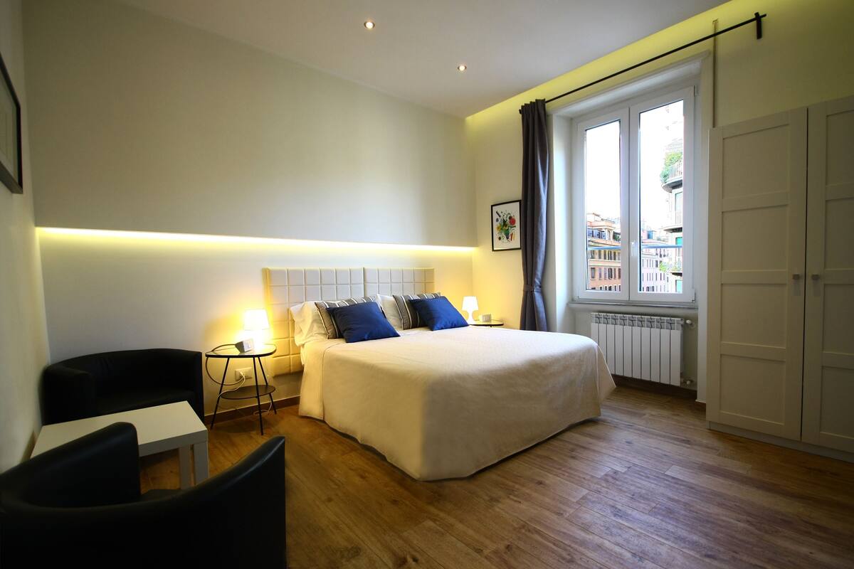 A hotel room with a bed, a small table, a chair, and a window with curtains.