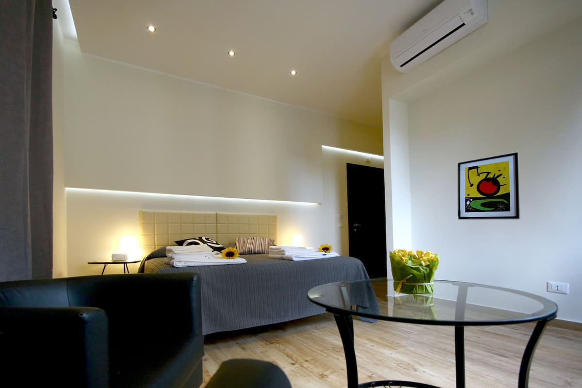 A modern hotel room with a bed, a glass table, a chair, and a wall-mounted air conditioner.