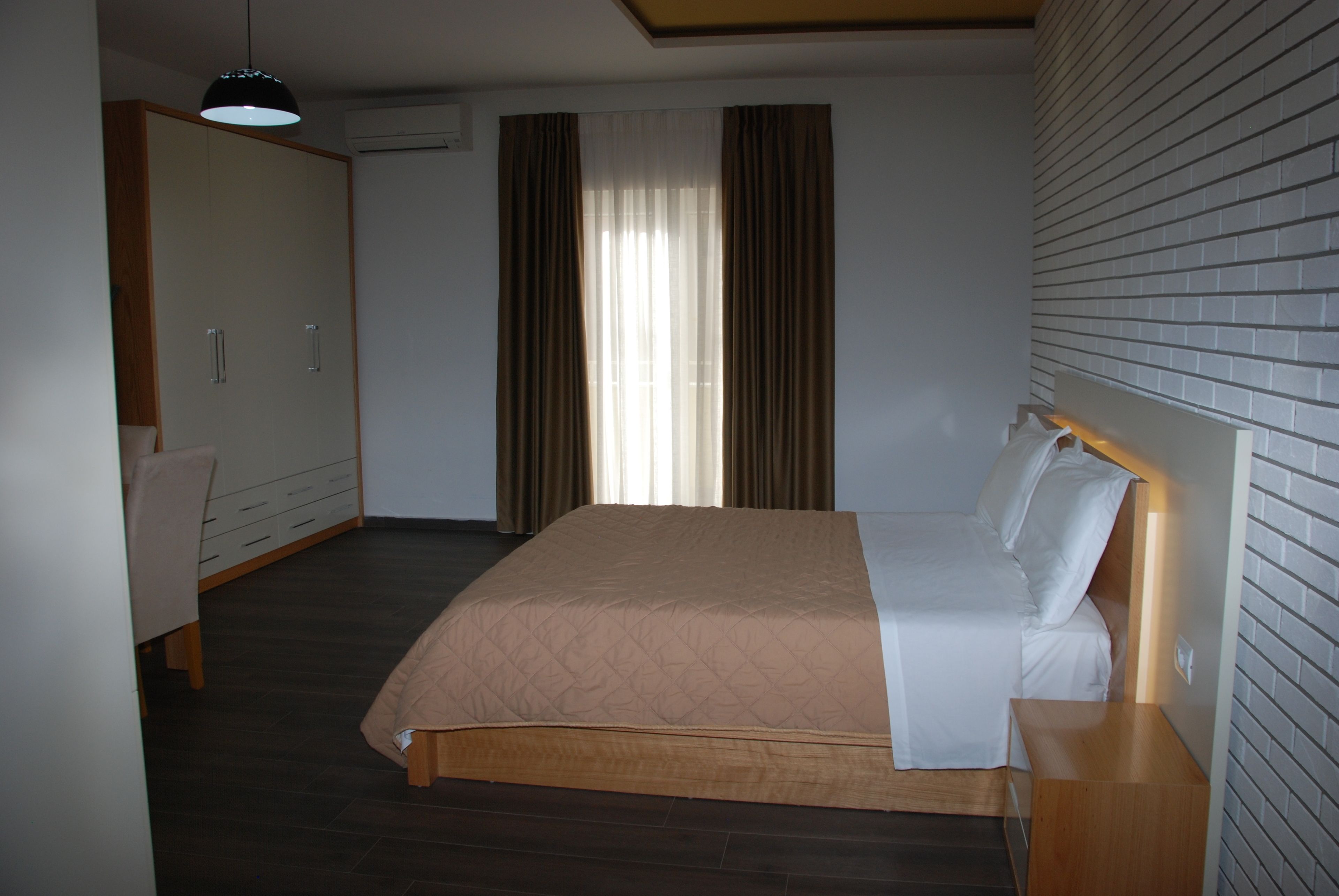 Quarto com cama, mesa de cabeceira de madeira, cadeira, guarda-roupa e janela com cortinas.