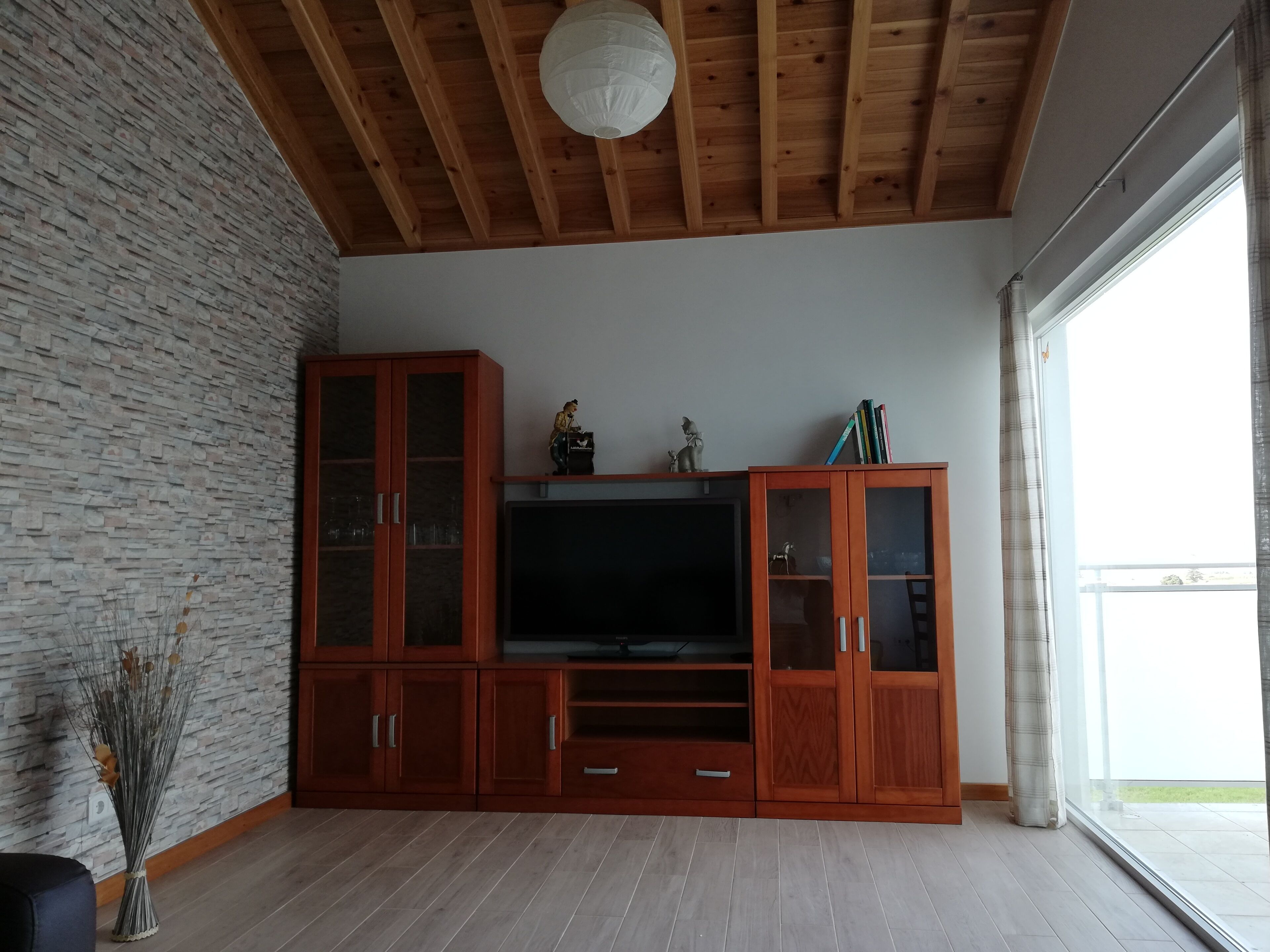 A living room with a stone wall, wooden ceiling beams, a large entertainment unit, and a view through a window.
