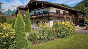 Un edificio tradicional de madera alpina con balcón, rodeado de jardines cuidados y montañas al fondo.