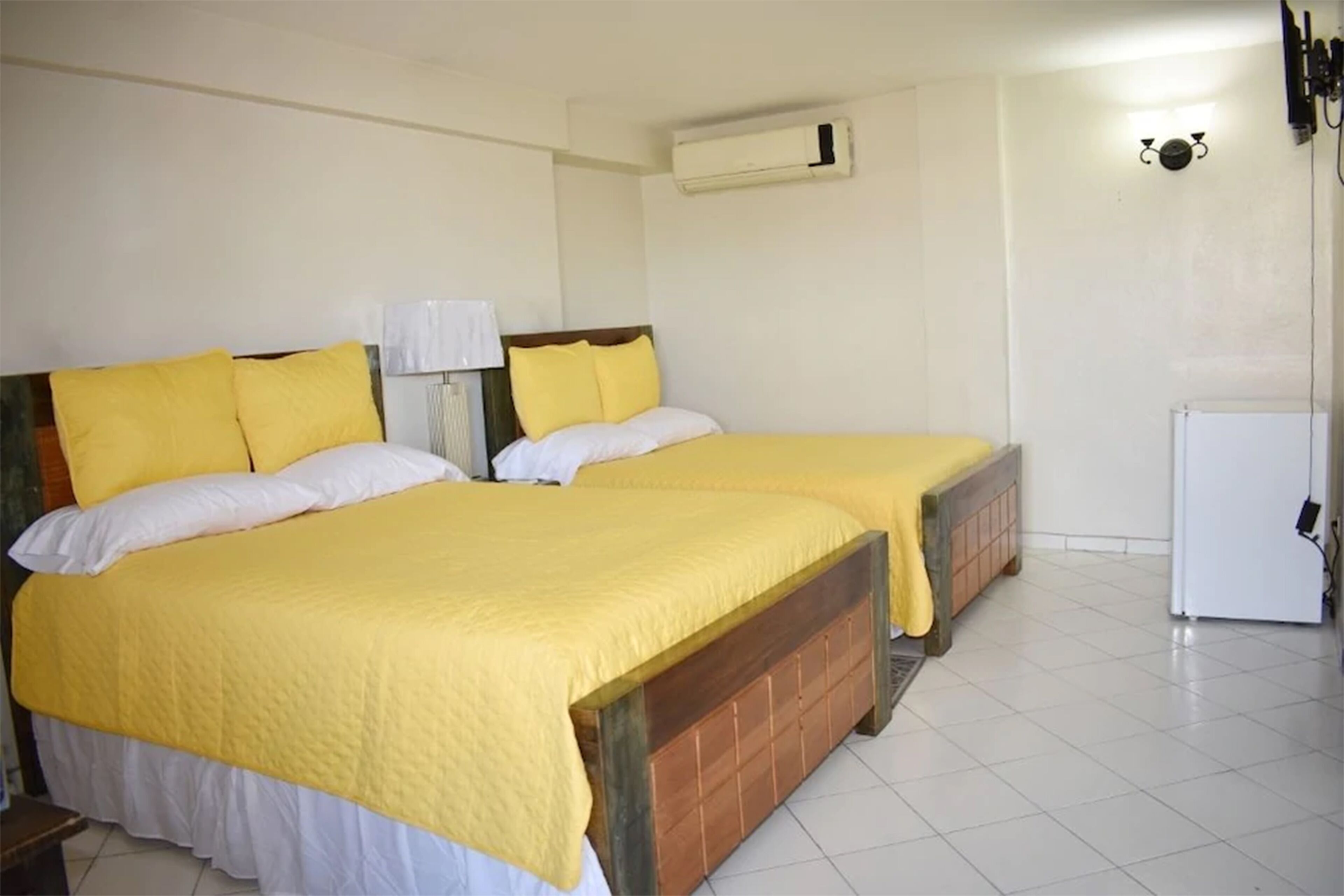 A room with two beds, each with a yellow bedspread and white pillows, a white radiator, and a white refrigerator.
