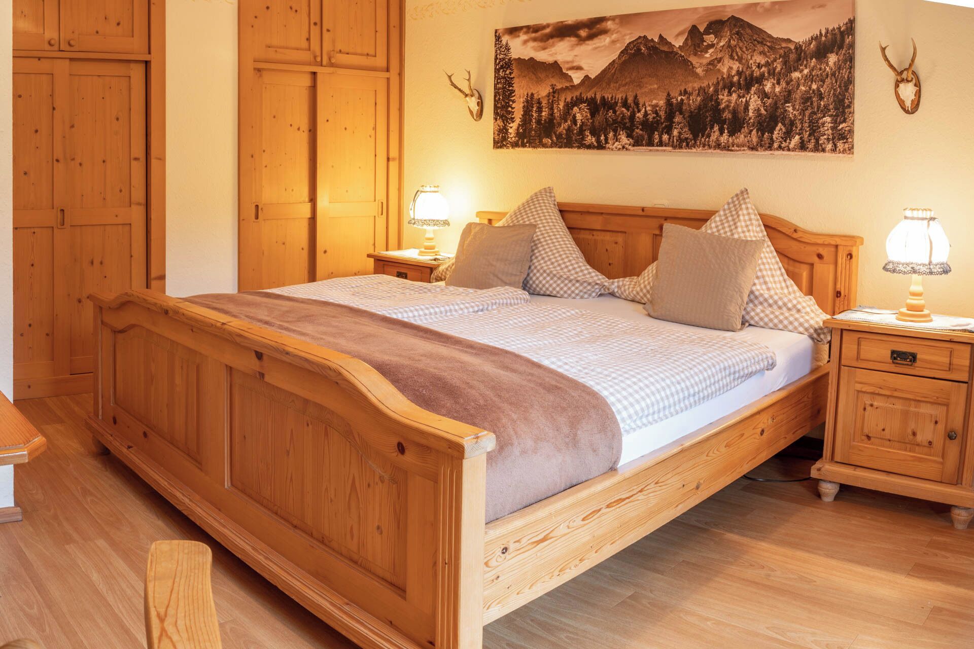 Un letto di legno con copripiumino a quadretti, due cuscini e una coperta. Due comodini con lampade sono presenti. Sopra il letto, un quadro raffigura un paesaggio montano.