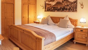 A wooden bed with a checkered bedspread, two pillows, and a blanket. There are two bedside tables with lamps. A mountain landscape painting is on the wall above the bed.