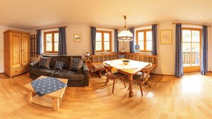 A spacious living room with a wooden floor, a sofa, a dining table with chairs, and a bench. There are large windows with curtains, a wooden cabinet, and a ceiling fan.