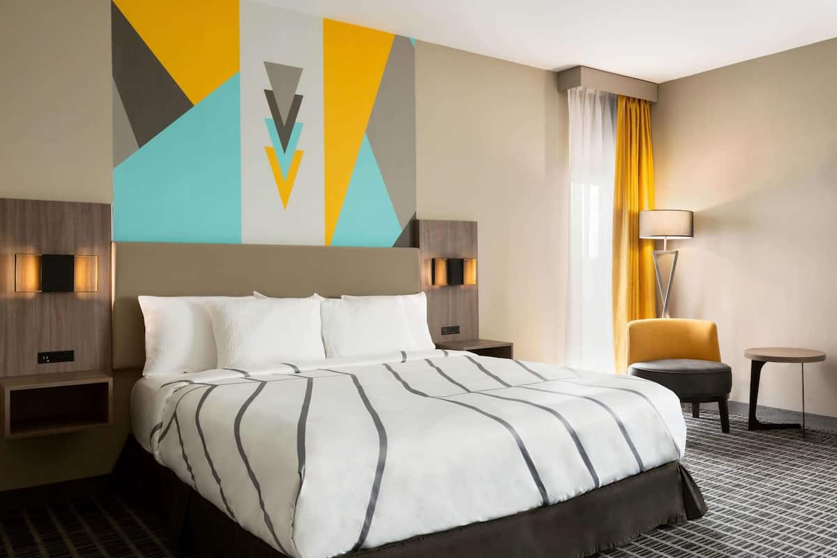 A hotel room with a large bed, a nightstand with a lamp, a chair, and a small table.
