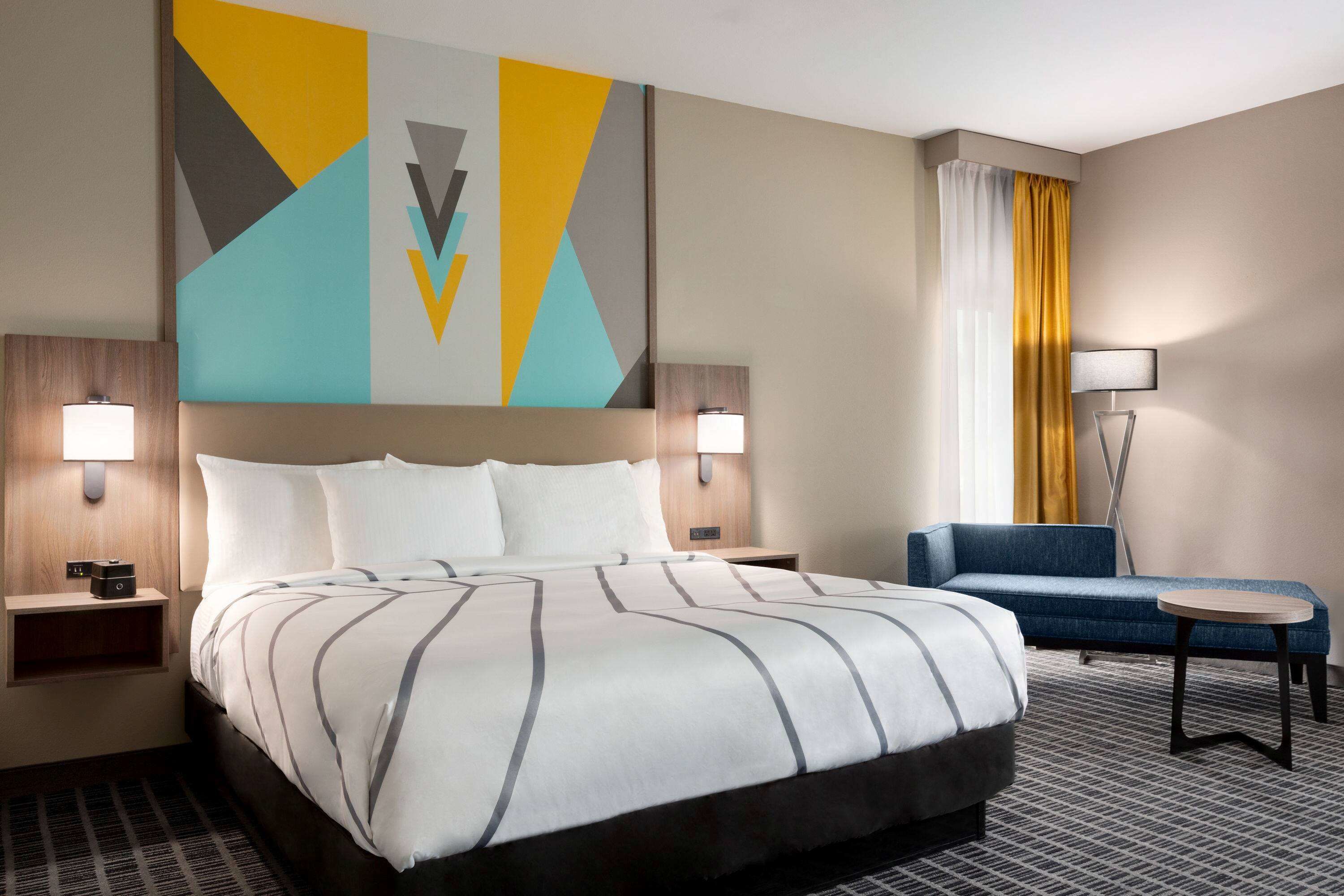 A hotel room with a large bed, two bedside tables, a desk, a chair, and a wall with geometric artwork.