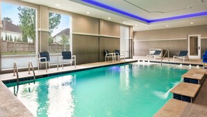 A swimming pool with clear blue water, surrounded by lounge chairs and tables, and large windows offering a view of the outside. - La Quinta Inn & Suites by Wyndham Richmond-Sugarland (Richmond)