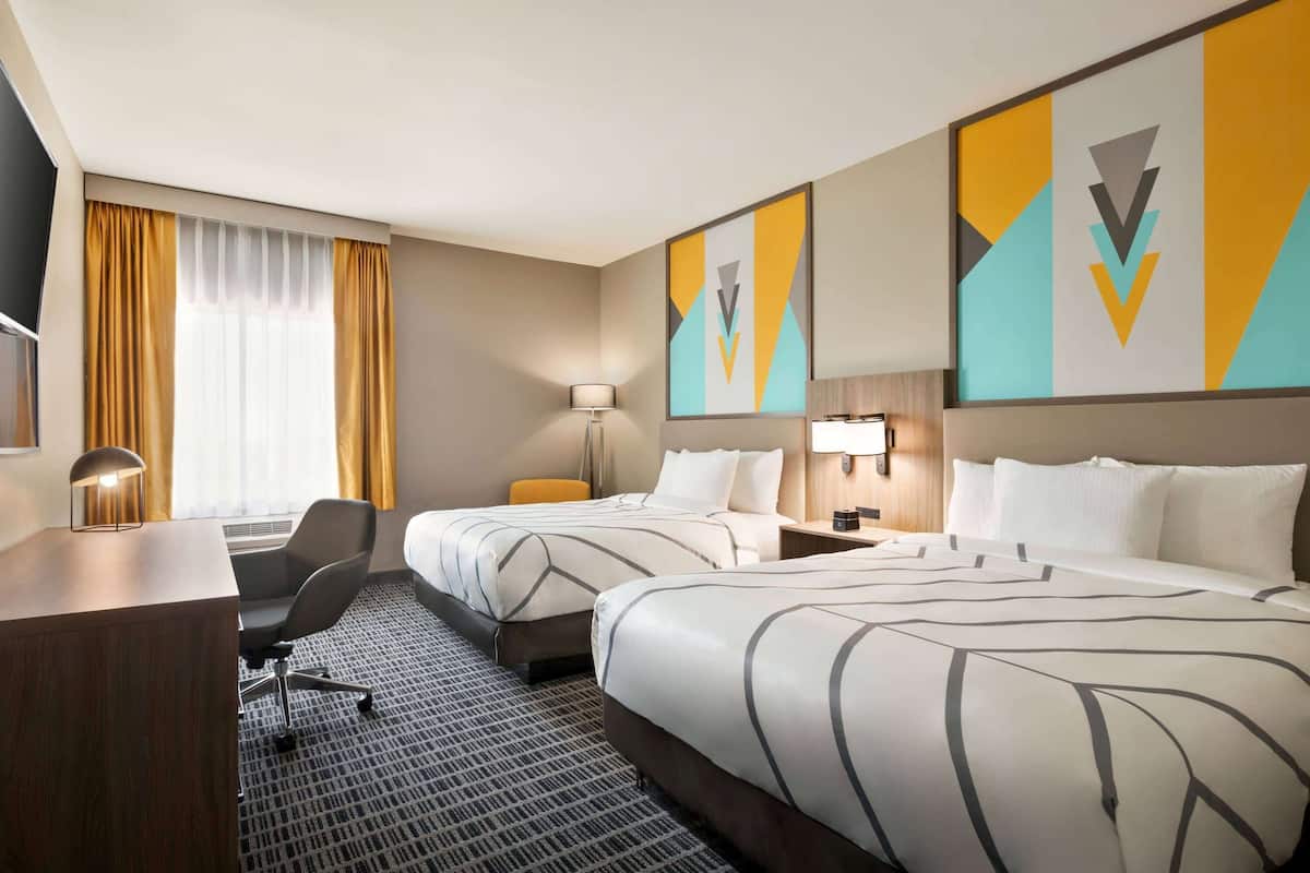 A hotel room with two beds, a desk, a chair, a lamp, and a TV.