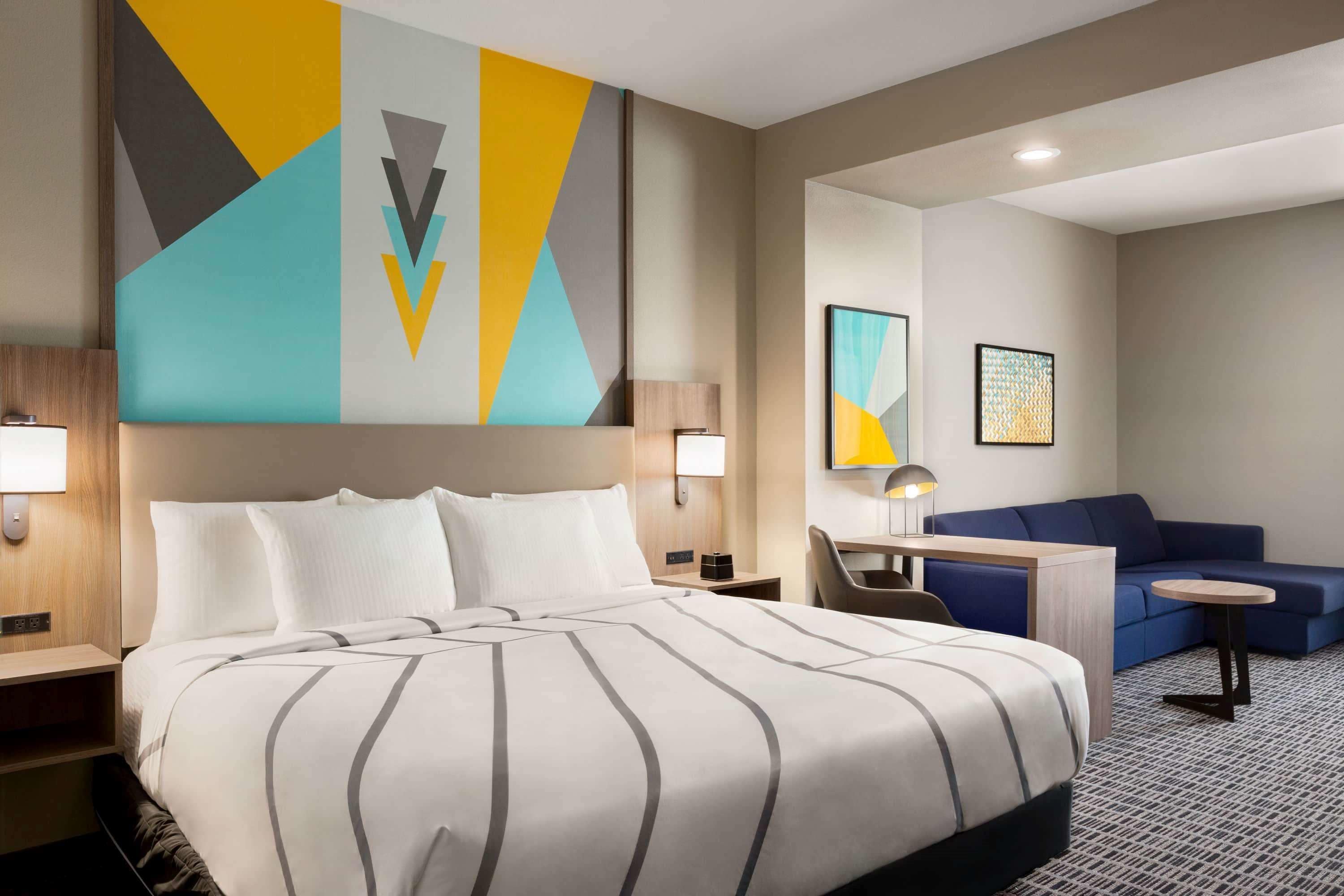 A hotel room with a large bed, a desk, a blue sofa, and a wall with geometric artwork.