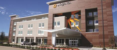 A modern hotel with a prominent 'La Quinta' sign, featuring a large sun logo. It has a flat roof, multiple windows, and a covered entrance area.
