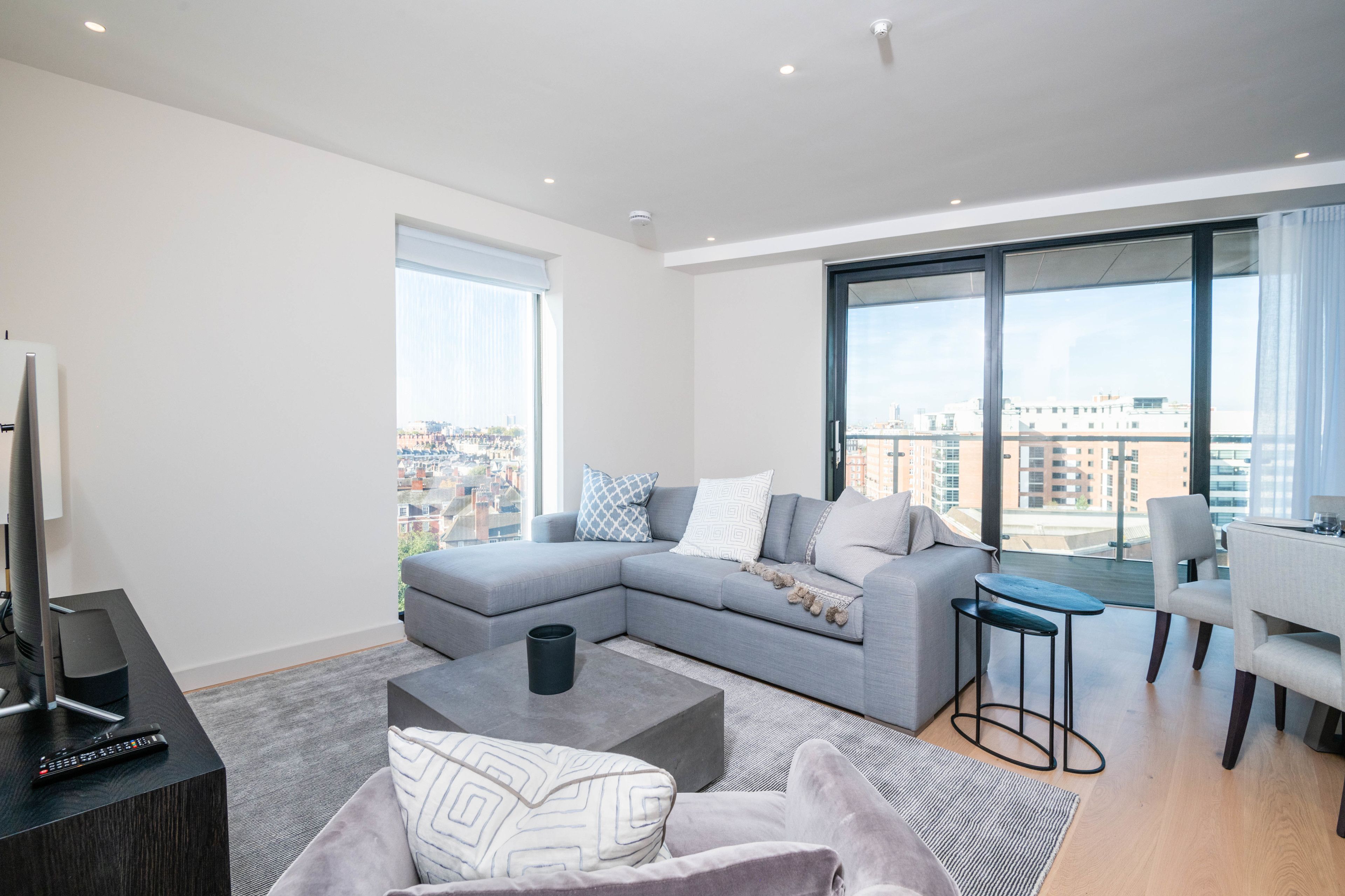 A modern living room with a grey sectional sofa, a coffee table, a TV, and a city view through large windows.