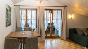 A dining area with a wooden table, four chairs, and a vase of flowers. There is a sofa and a wall-mounted light fixture. - Vacation apartment 7 for up to 4 people, with 2 extra bedrooms and kitchen in the living/dining area (Reit im Winkl)
