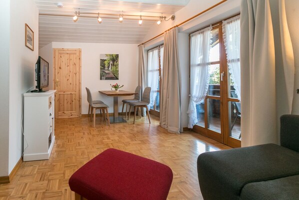 Une pièce dotée d’un plancher en bois, d’une table à manger avec des chaises, d’un pouf rouge, d’un canapé gris et d’une porte-fenêtre coulissante donnant sur un espace extérieur.