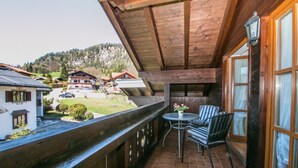 A balcony with a wooden ceiling, a small table with chairs, and a view of a village with houses and a mountain in the background. - Vacation apartment 7 for up to 4 people, with 2 extra bedrooms and kitchen in the living/dining area (Reit im Winkl)
