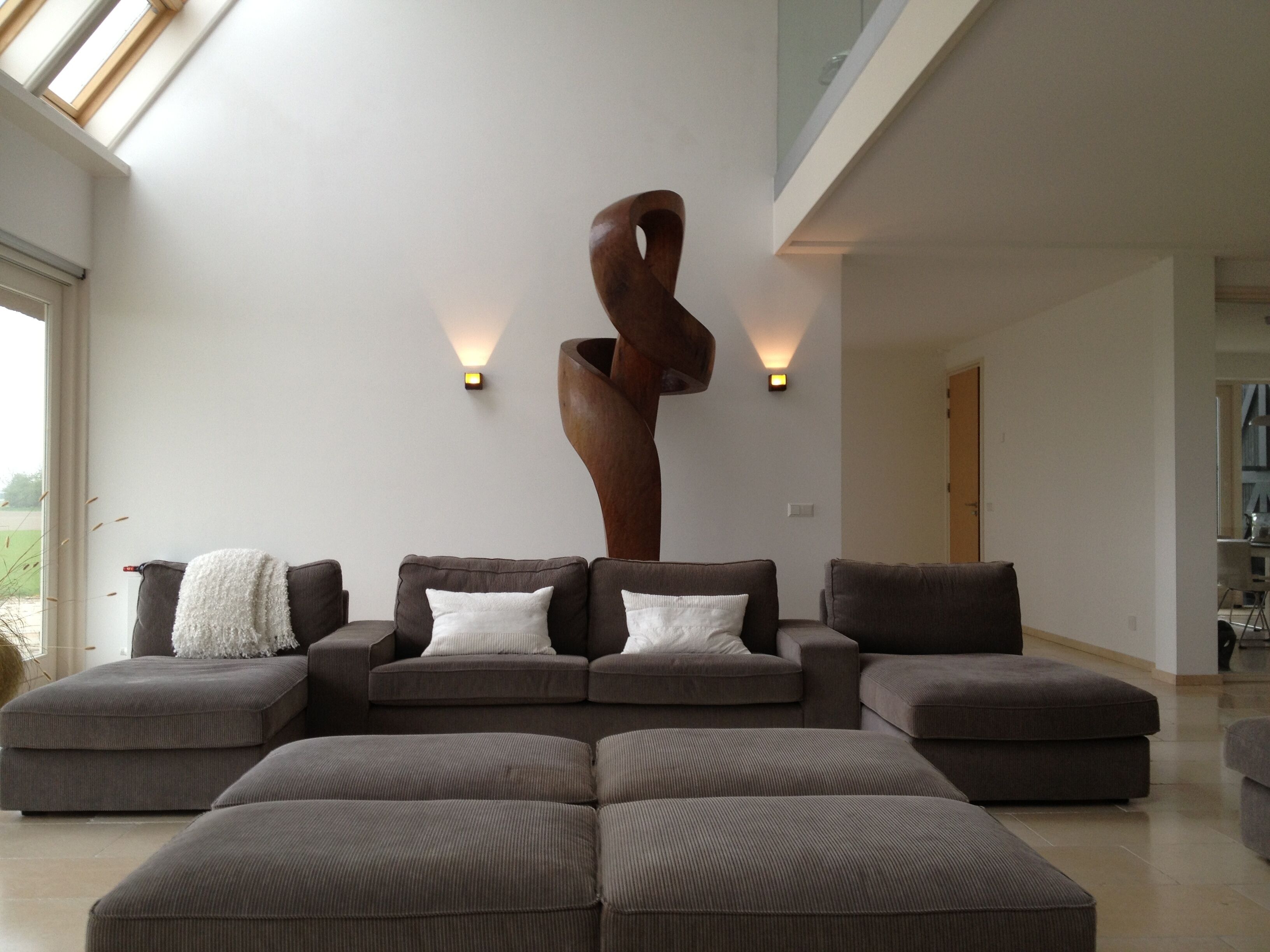A modern living room with a sectional sofa, a large wooden sculpture, and two wall-mounted lights.