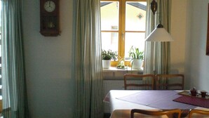 A dining area with a table, chairs, a clock, curtains, and a window with plants.