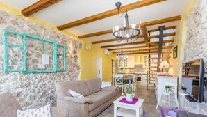 A cozy living room with a stone wall, wooden beams, a chandelier, a sofa, a coffee table, and a kitchen in the background. - Rustic holiday house KATE (Čižići)