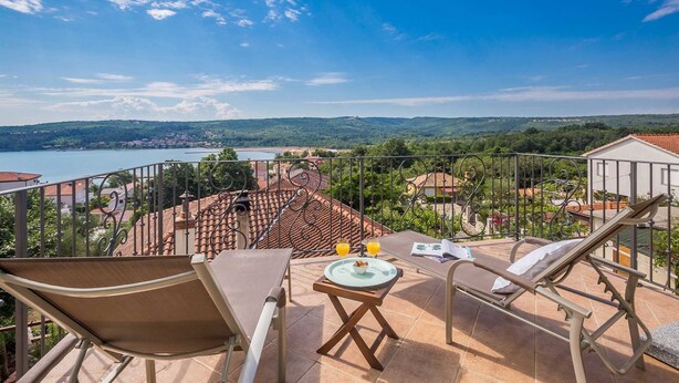 A balcony with two lounge chairs, a small table with a tray and two glasses of orange juice, and a scenic view of a lake and surrounding hills. - Rustic holiday house KATE (Čižići)