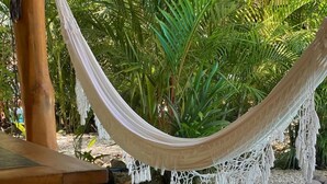 Una hamaca atada entre postes de madera, una mesa de madera con cojines amarillos y una piscina rodeada de follaje.