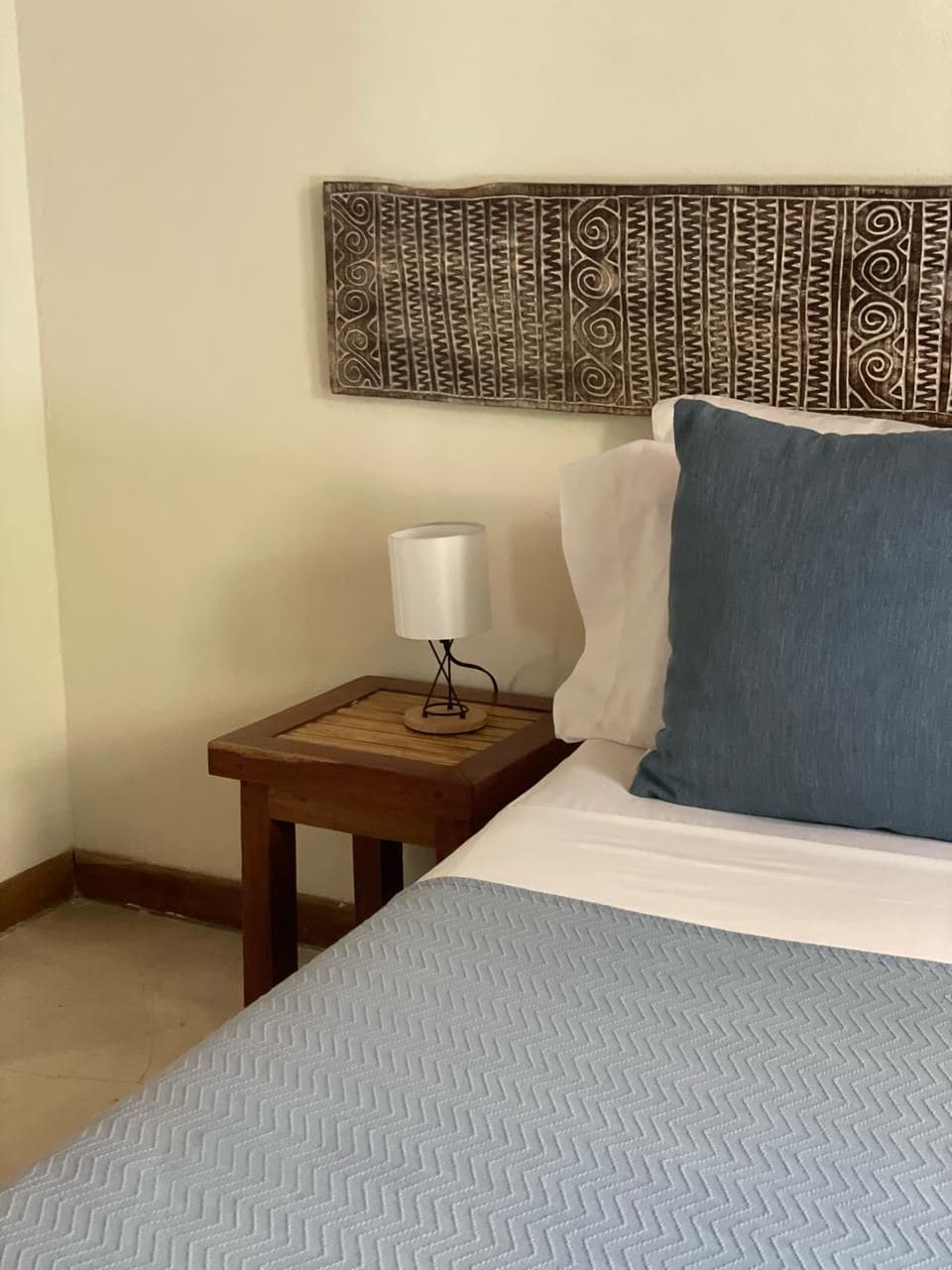 A neatly made bed with a blue pillow and a white pillow, a small wooden nightstand with a lamp, and a decorative wall piece above the bed.