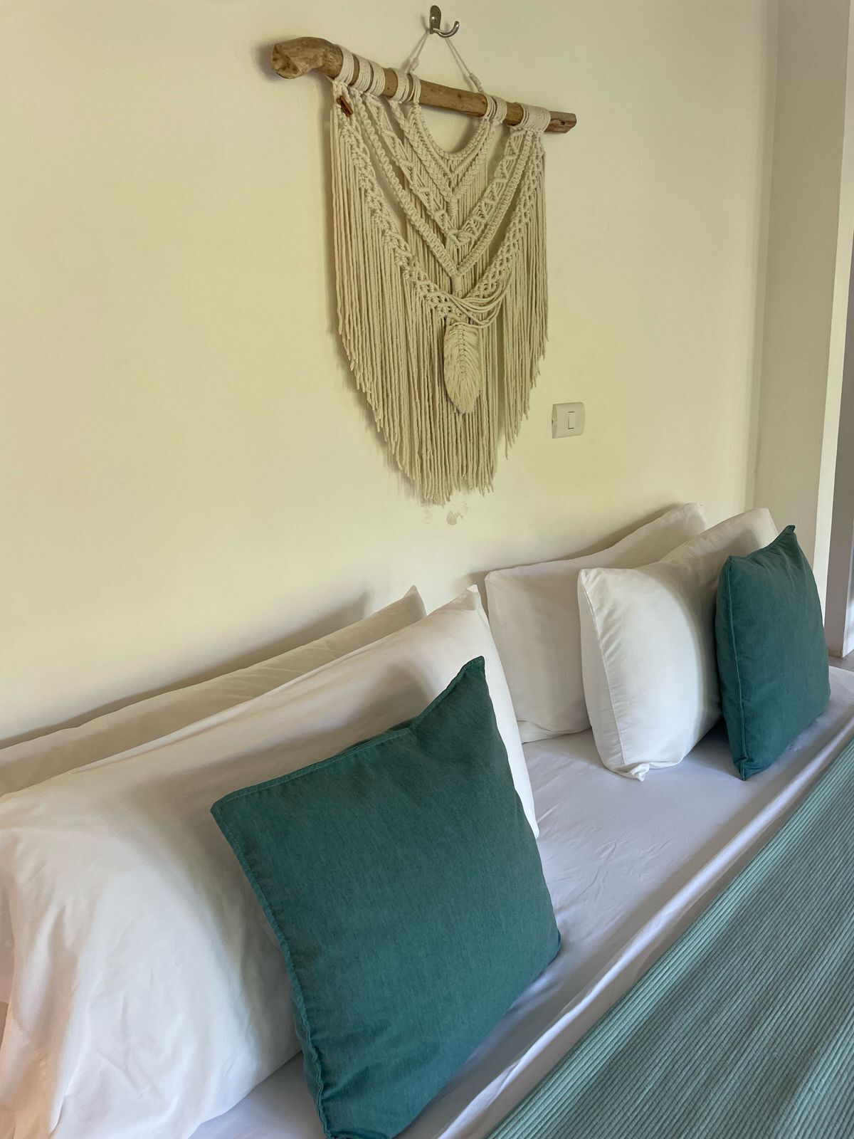 Un lit avec des oreillers blancs et verts, une décoration murale en macramé et une tête de lit en bois.