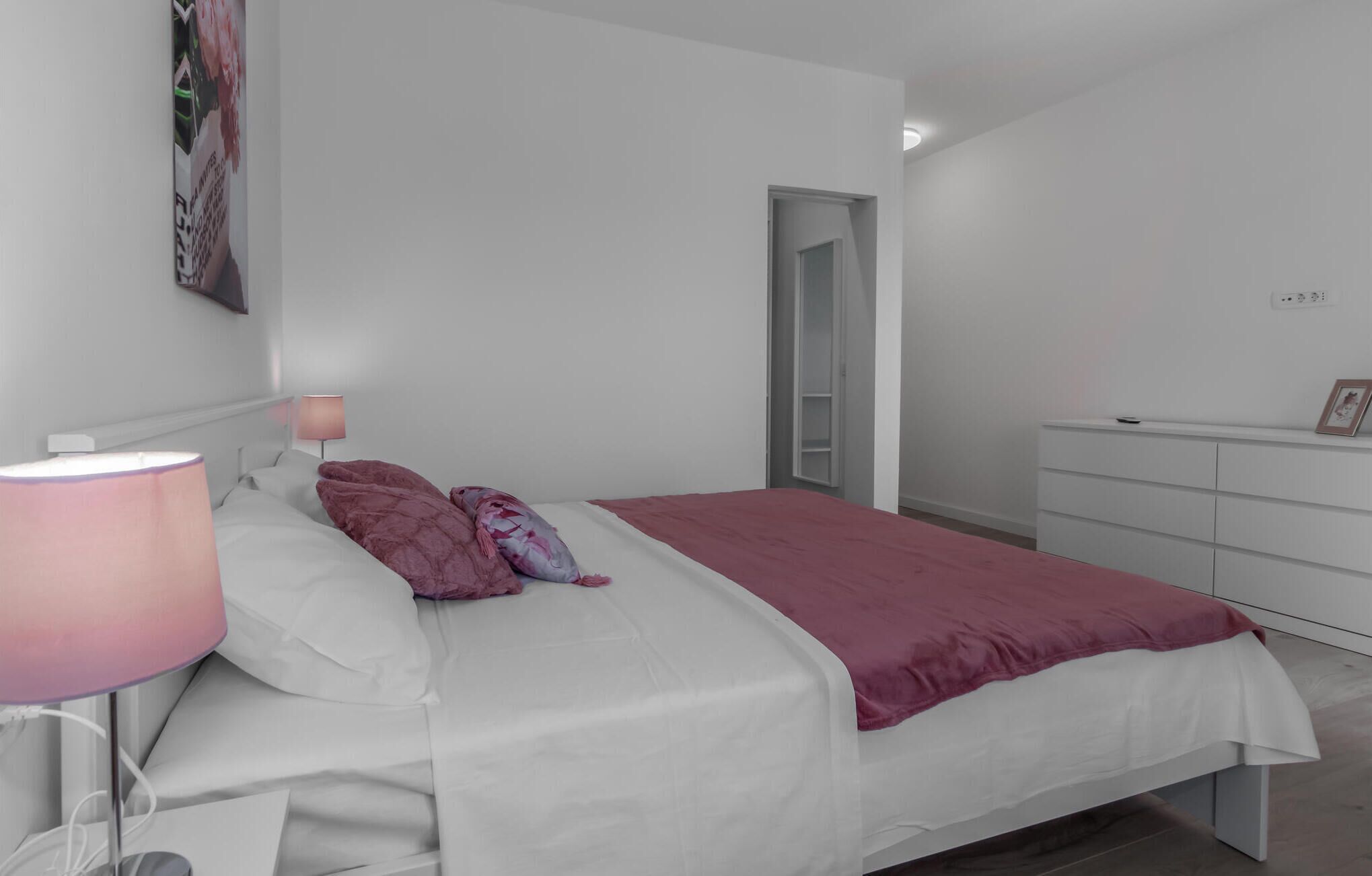 A bedroom with a bed, a bedside lamp, a dresser, and a door leading to another room.
