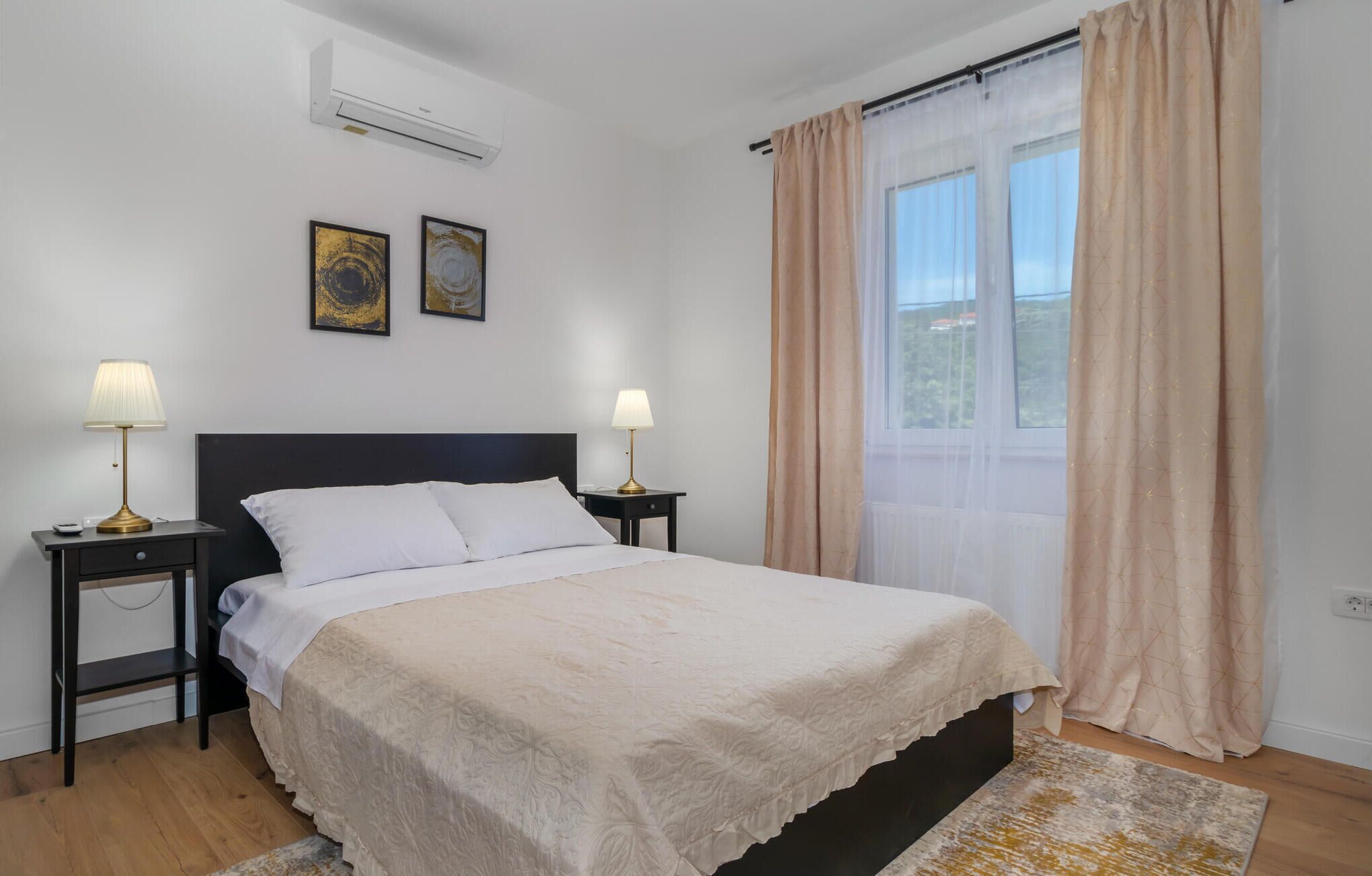 Une chambre à coucher avec un lit, deux tables de chevet avec des lampes, une fenêtre avec des rideaux et deux tableaux encadrés accrochés au mur.