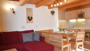 A cozy living area with a red sofa, wooden dining table, and chairs. A kitchen with wooden cabinets and a microwave is visible in the background. - Ferienhaus Gänseblümchen (Doppelhaushälfte) 65 qm (Höslwang)