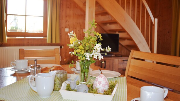 A wooden dining table with a floral centerpiece, white cups, a glass pitcher, and a plate of pastries. - Ferienhaus Gänseblümchen (Doppelhaushälfte) 65 qm (Höslwang)