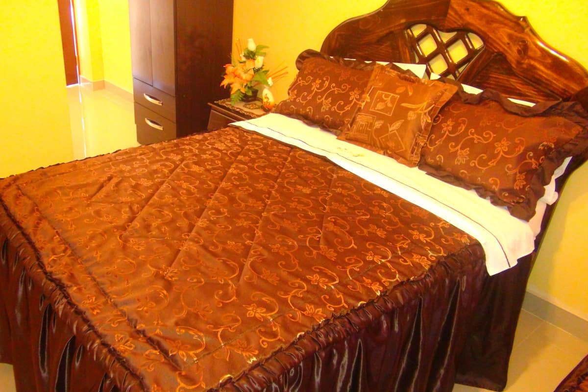 A bed with a brown quilt and matching pillows, a wooden headboard, and a nightstand with a vase of flowers.